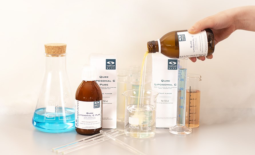 Product packaging, bottles and measuring glasses on a table. A hand pours a bottle of QURE Liposomal C into a measuring glass with water.